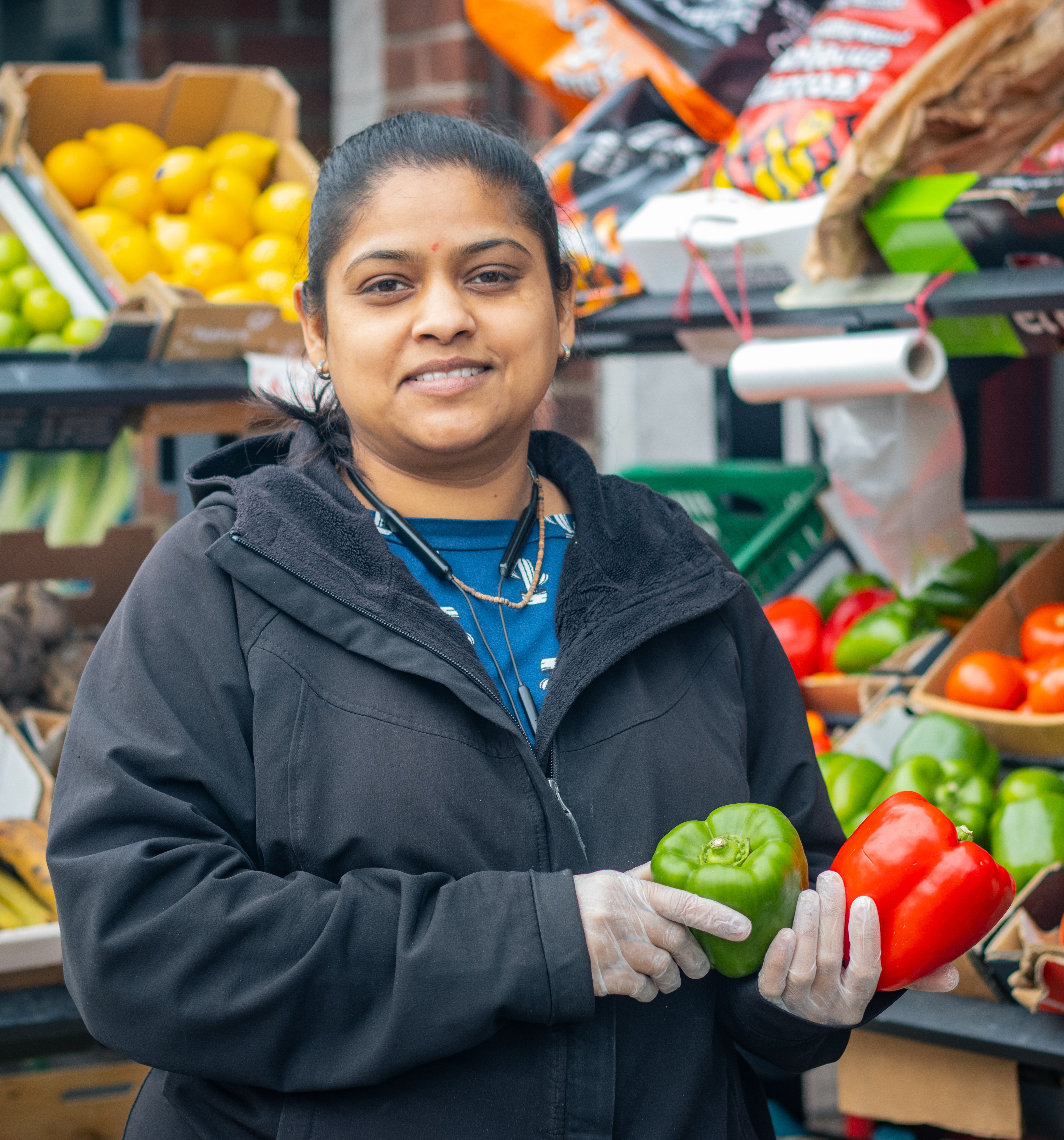 A woman stands outside a shop selling fruit and vegetables holding two peppers